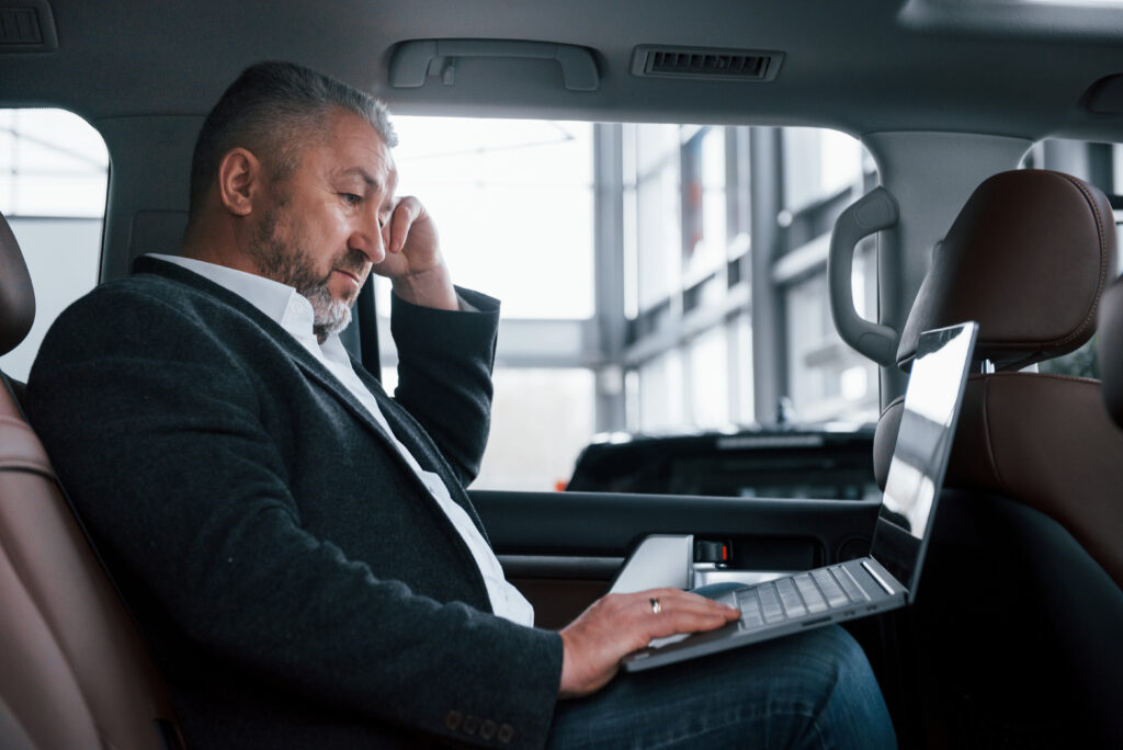 Side view. Working on a back of car using silver colored laptop. Senior businessman.